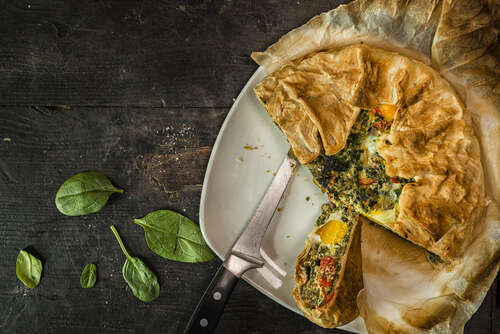 an above view of a torta pasqualina on a white plate on a dark wooden table with some basil leaves and a kitchen knife