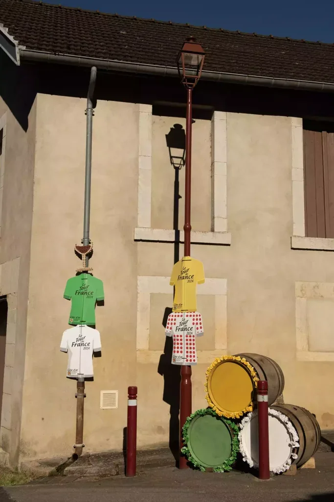 A gutter and lamppost in front of a stone house decorated with four cardboard signs in the shape of Tour de France jerseys, one in white, one in green, one in yellow and one with red spots on white and a stack of three barrels with the ends painted in yellow, green and white