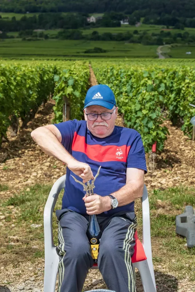 A man in a blue French national team football t-shirt with a red stripe and blue Adidas tracksuit trousers and cap sitting on a plastic chair in a vineyard opening a bottle of wine between his knees with a corkscrew