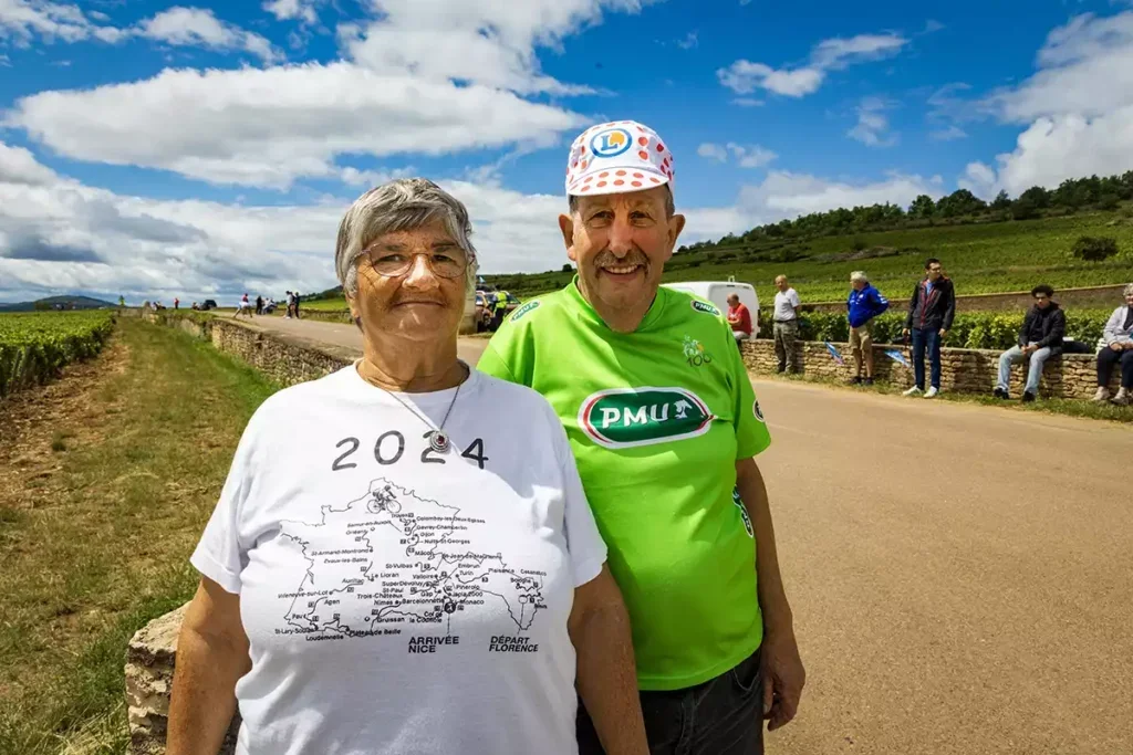 An older man in a red-spotted white cycling cap and a bright green cycling shirt with a sponsor's sign for PMU and an older woman in a white t-shirt with the 2024 Tour de France route on the front stand by a road through a vineyard during the 2024 Tour de France.